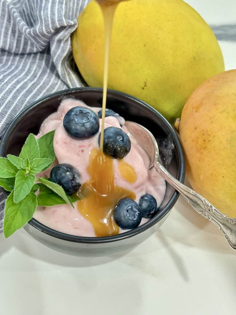 caramel sauce being poured over black bowl with silver spoon filled with raspberry ice cream topped with blueberries and herb in front of striped napkin and two ripe mangoes