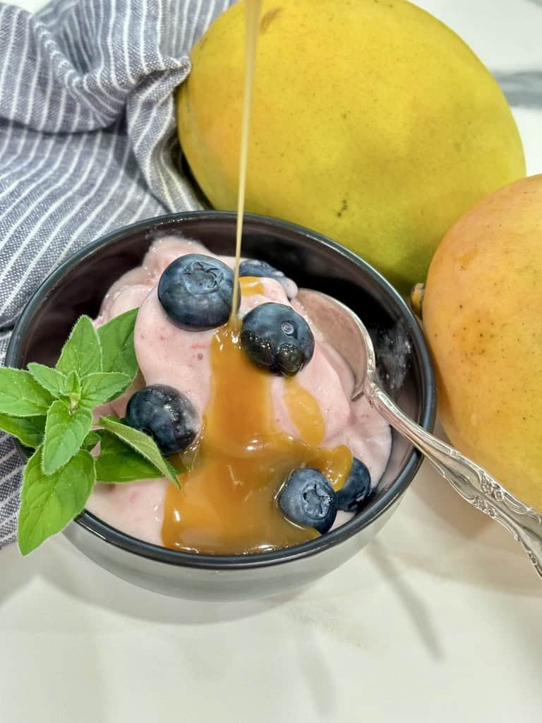 caramel sauce being poured over black bowl with silver spoon filled with raspberry ice cream topped with blueberries and herb in front of striped napkin and two ripe mangoes