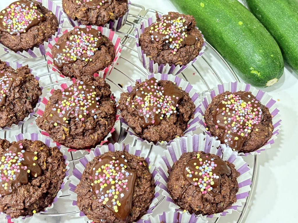 Cooling rack of chocolate muffins topped with chocolate and sprinkles, next to two zucchini