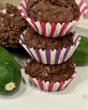 Stack of 3 chocolate zucchini muffins next to fresh zucchini