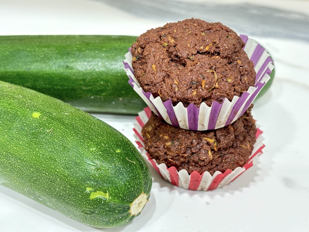 Two dark chocolate zucchini muffins next to two zucchinis