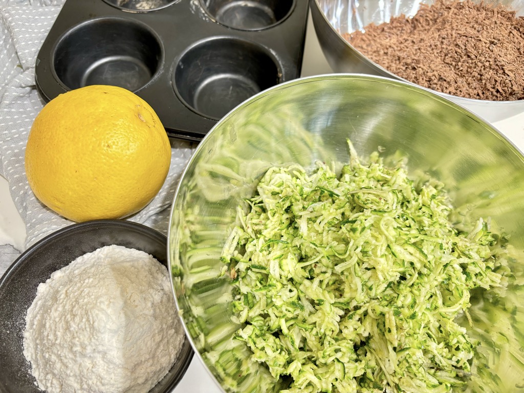 Grated zucchini, grated chocolate, lemon and flour next to muffin tin