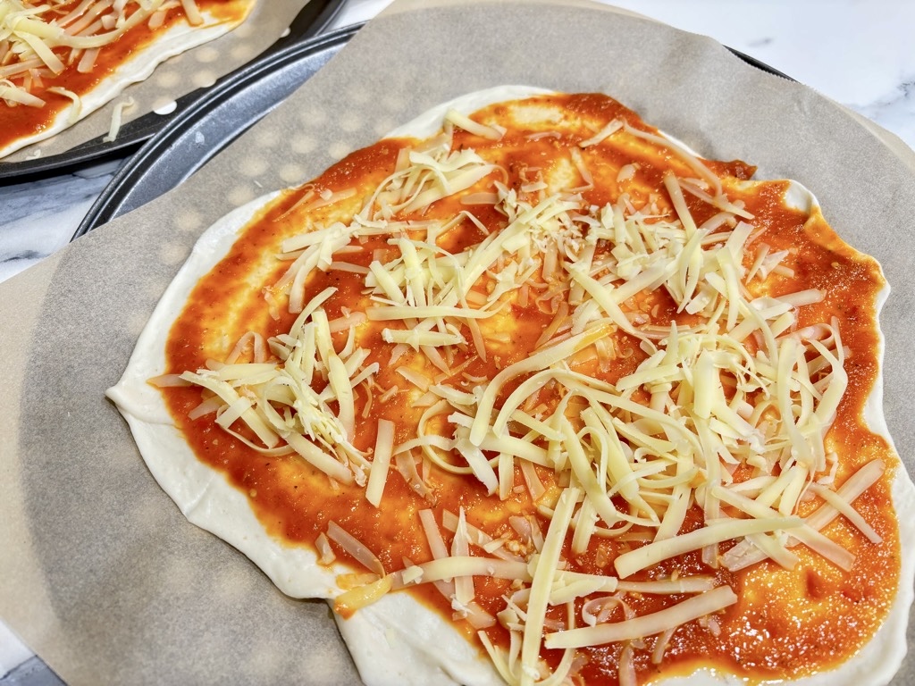 raw pizza dough on parchment paper lined baking tray, topped with tomato sauce and shredded cheese