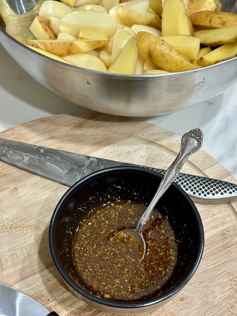 Silver bowl filled with raw potato wedges next to wooden board with small bowl of marinade standing on top of it