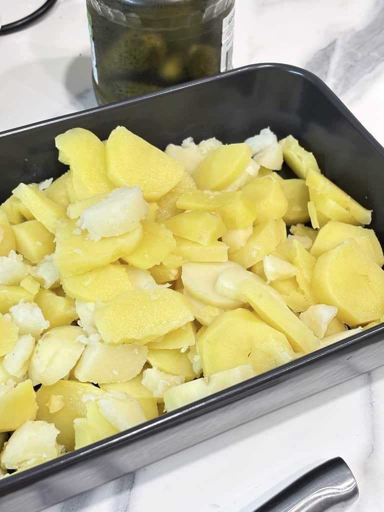 Black shallow dish of sliced potatoes standing on kitchen counter in front of pickle jar