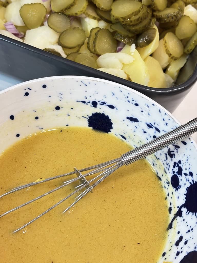 Small bowl of salad dressing in front of potato salad bowl