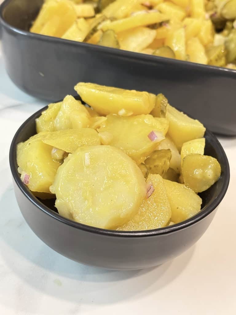 Small black bowl of potato salad standing in front of black shallow dish of potato salad on kitchen counter