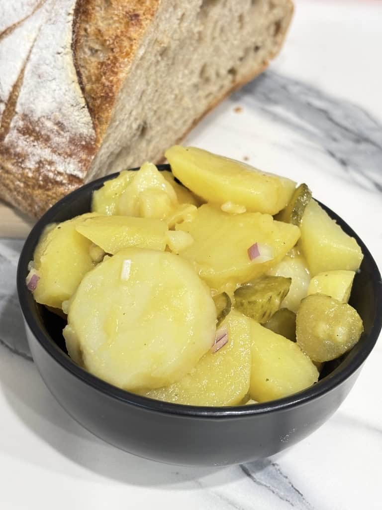 Small black bowl of potato salad standing on kitchen counter in front of sliced rustic bread