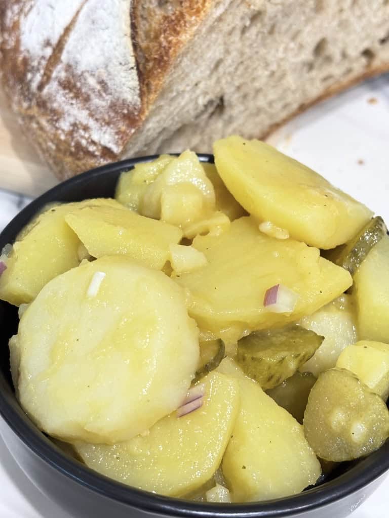 Small black bowl of potato salad standing on kitchen counter in front of rustic bread loaf