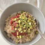 Rice Flakes Porridge in grey bowl with silver spoon, topped with rhubarb and chopped pistachios on white linen napkin