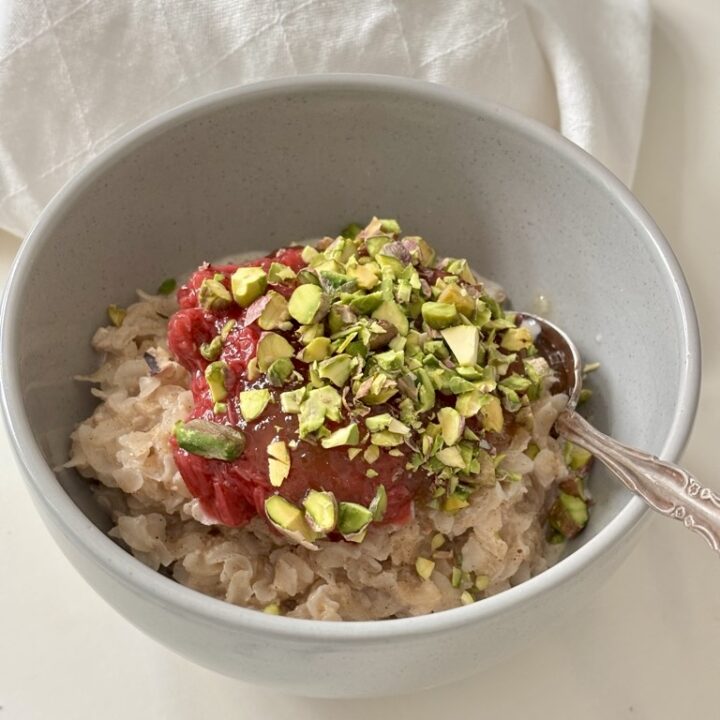 Rice Flakes Porridge in grey bowl with silver spoon, topped with rhubarb and chopped pistachios on white linen napkin