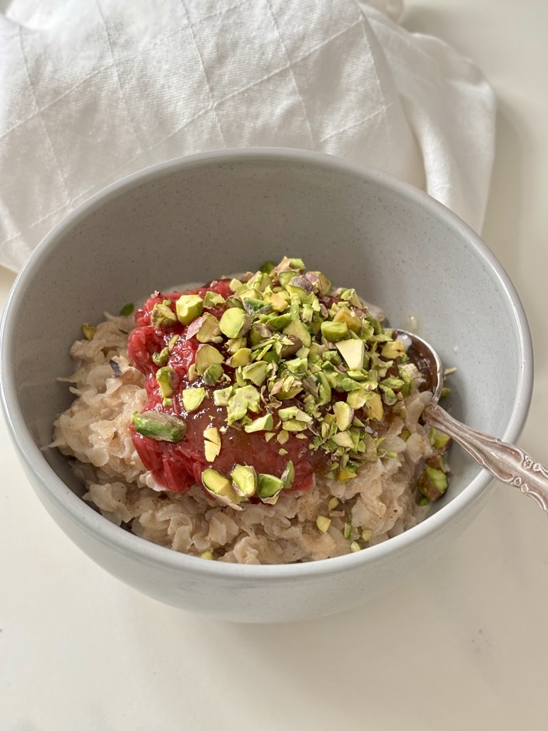 Rice Flakes Porridge in grey bowl with silver spoon, topped with rhubarb and chopped pistachios on white linen napkin