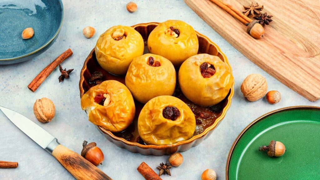 Baked stuffed apples in serving bowl, next to nuts, spices, platters and wooden tray