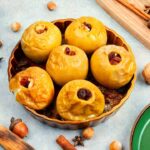 Baked stuffed apples in serving bowl, next to nuts, spices, platters and wooden tray