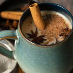 Ceramic mug filled with chai latte topped with spices, next to strainer and spices on table