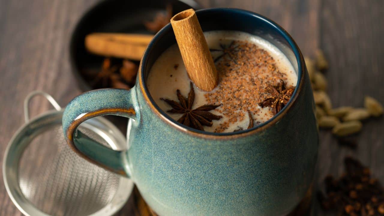 Ceramic mug filled with chai latte topped with spices, next to strainer and spices on table