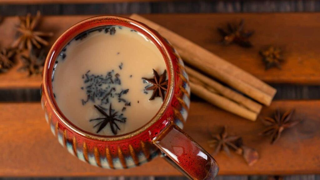 Red Ceramic mug filled with chai latte topped with spices, next to cinnamon sticks and star anise on table