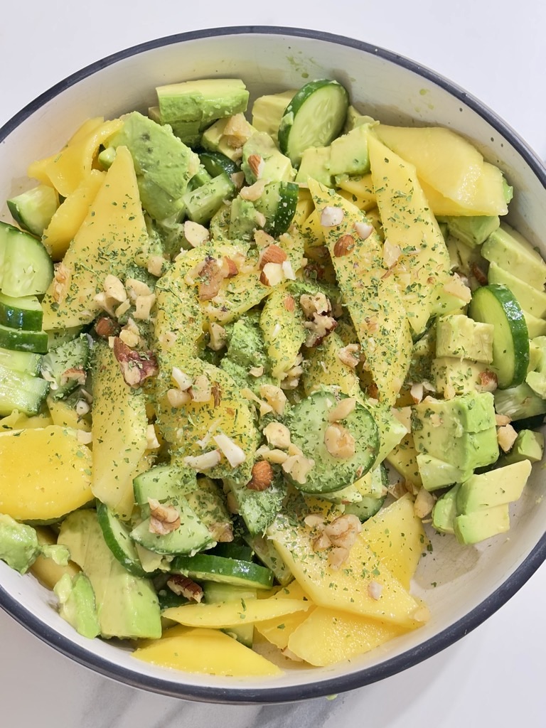 Top down shot of Mango, cucumber and avocado salad in cream bowl, topped with greens and chopped nuts