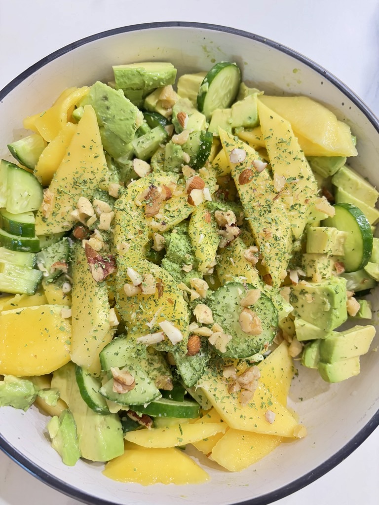 Mango, cucumber and avocado salad in cream bowl, topped with greens and chopped nuts on white surface