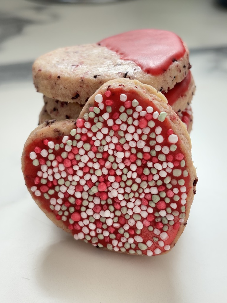 Stack of heart shaped cookies with single cookie covered in red icing and sprinkles leaning against it