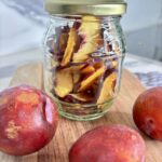 Glass jar filled with dehydrated plums on wooden board, surrounded by three whole plums