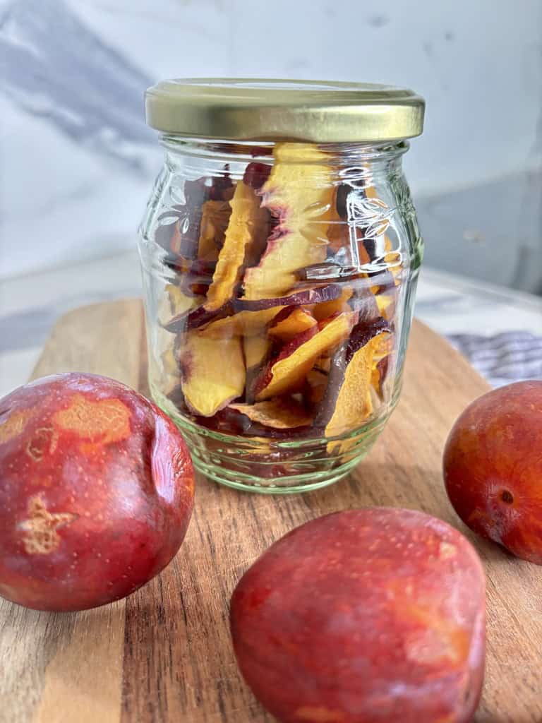 Glass jar filled with dehydrated plums on wooden board, surrounded by three whole plums