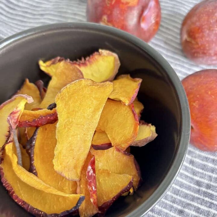 Dehydrated plum slices in black bowl on striped linen cloth, in front of four whole plums