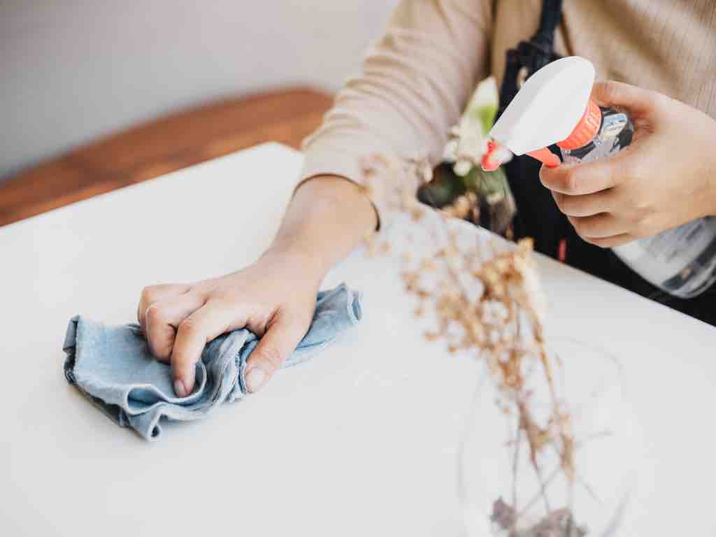 Close-up of a person cleaning a white surface using a DIY all-purpose cleaner in a spray bottle and a blue cloth, promoting natural home cleaning solutions.