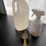 A jug of clear homemade all-purpose cleaner, a white spray bottle, and a measuring cup placed on a black countertop in natural light—ideal for a DIY cleaning recipe tutorial.