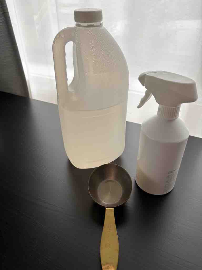 A jug of clear homemade all-purpose cleaner, a white spray bottle, and a measuring cup placed on a black countertop in natural light—ideal for a DIY cleaning recipe tutorial.