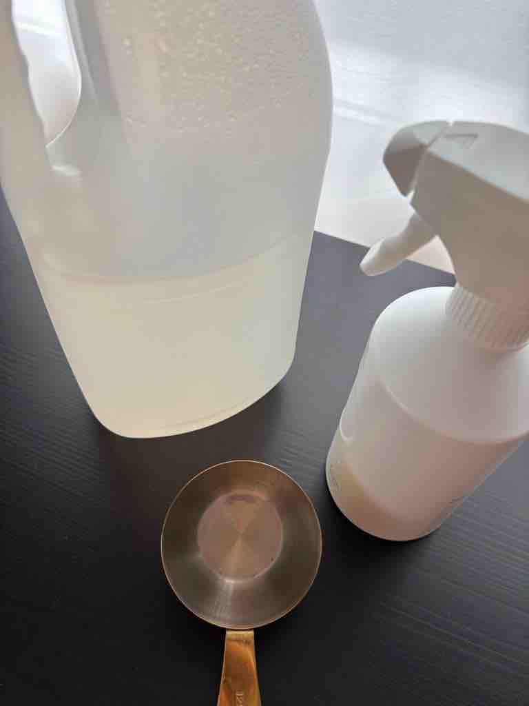 Top-down view of a vinegar-based DIY all-purpose cleaner setup featuring a spray bottle and a brass measuring cup on a sleek black surface.