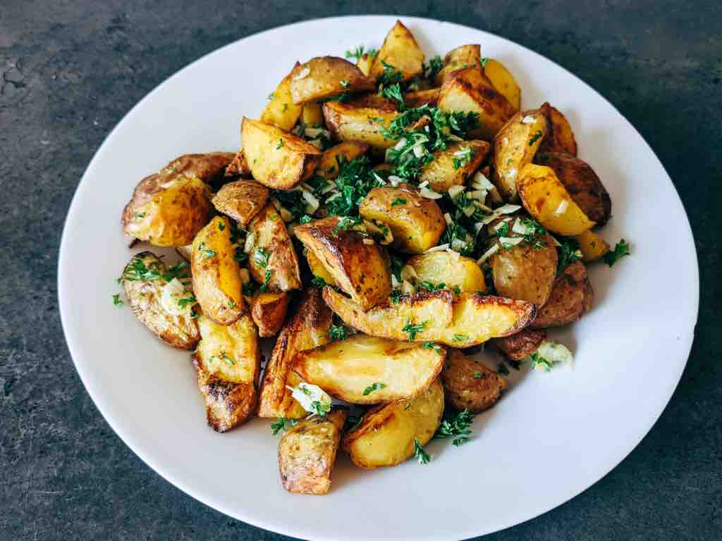 A white plate filled with golden, crispy roasted potato wedges garnished with chopped parsley and minced garlic on a dark grey surface.
