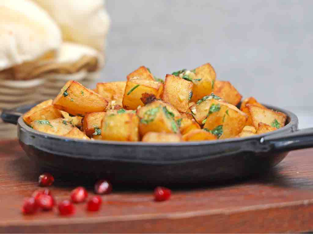 A black skillet filled with cubed, pan-fried golden potatoes mixed with fresh herbs, set on a wooden board with scattered pomegranate seeds and flatbread in the background.