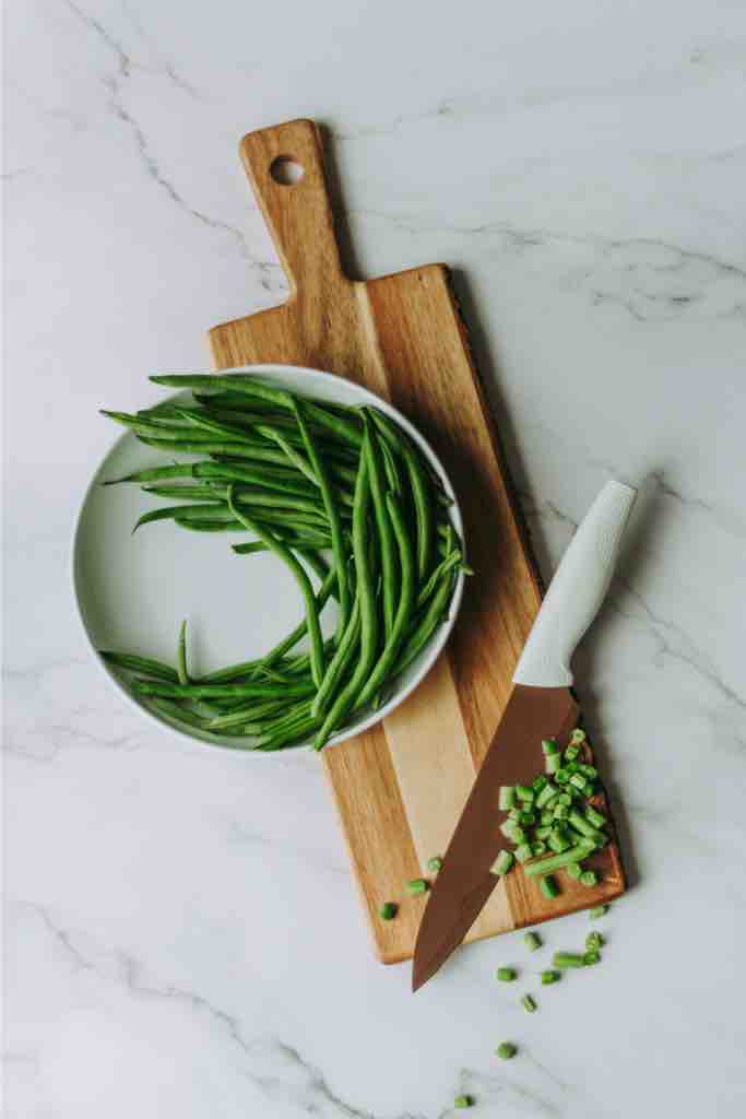A bowl of fresh green beans resting on a rustic wooden chopping board beside a sharp kitchen knife with chopped beans, set on a clean marble countertop—perfect prep for a hearty homemade meal.