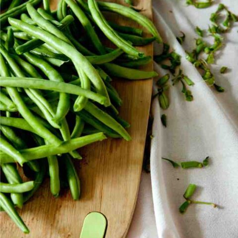 A generous handful of trimmed green beans on a wooden chopping board with scattered trimmings on a linen cloth—messy, honest, and beautifully real kitchen prep at its best.