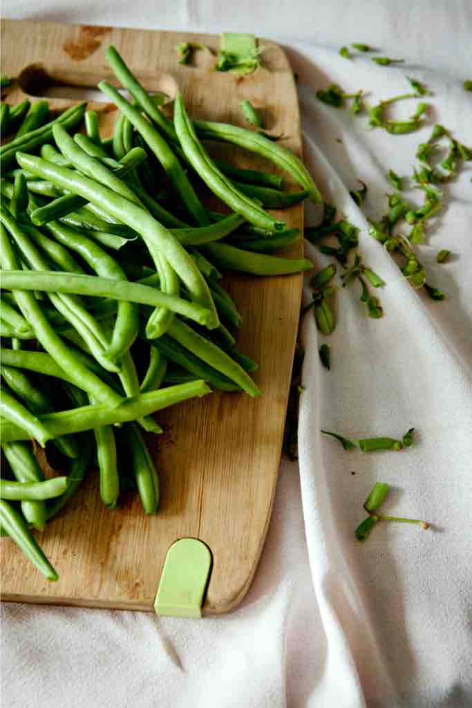 A generous handful of trimmed green beans on a wooden chopping board with scattered trimmings on a linen cloth—messy, honest, and beautifully real kitchen prep at its best.