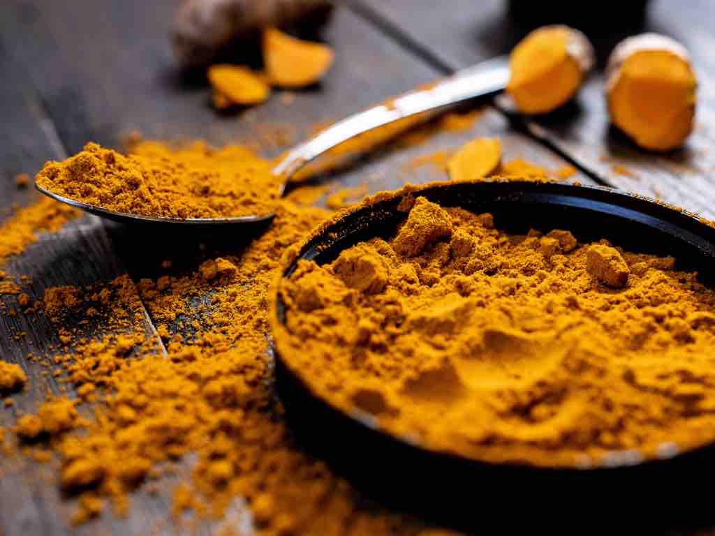 Close-up of vibrant turmeric powder in a black bowl and metal spoon, surrounded by scattered turmeric on a rustic wooden surface—showcasing the rich golden color and earthy texture of this powerful spice.