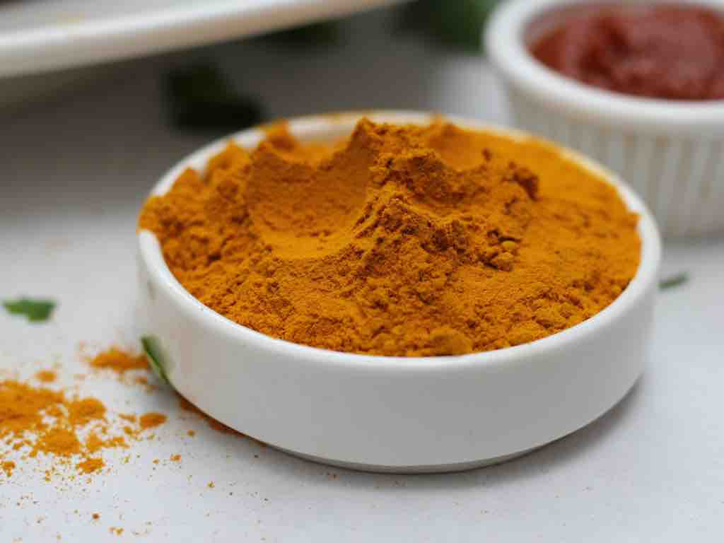 Small white bowl filled with heaped turmeric powder, surrounded by light turmeric dust and herbs—perfect for illustrating turmeric’s use in both culinary dishes and homemade remedies.