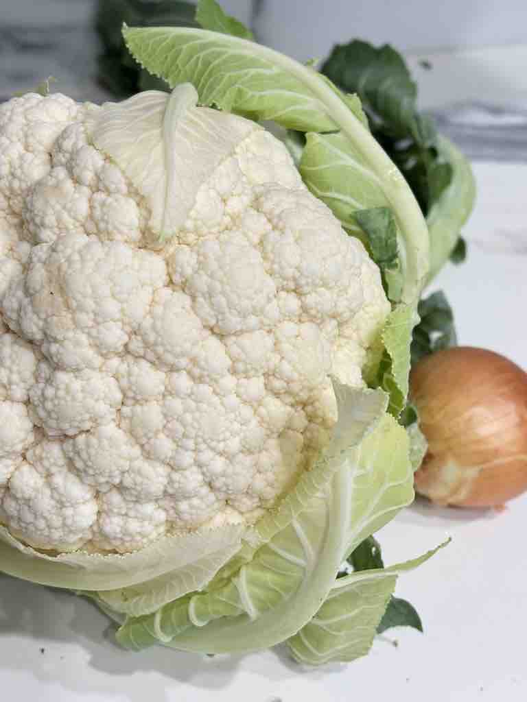 Fresh head of cauliflower with leaves and a brown onion on a marble countertop – perfect base for homemade cauliflower soup.