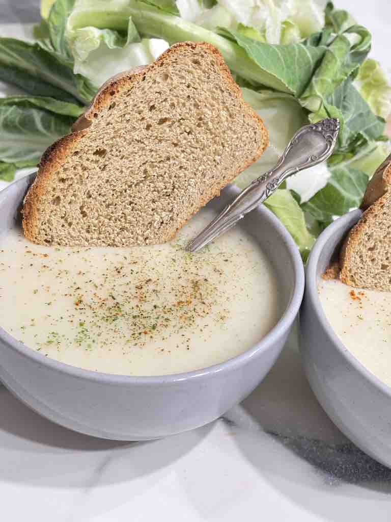 Thick slice of country-style bread with creamy cauliflower soup, surrounded by fresh cauliflower leaves.