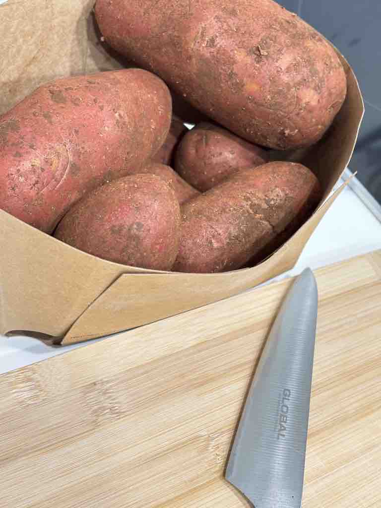 A rustic paper tray filled with unwashed red potatoes beside a cutting board and knife, about to be prepped for a hearty potato bake or classic potato gratin.