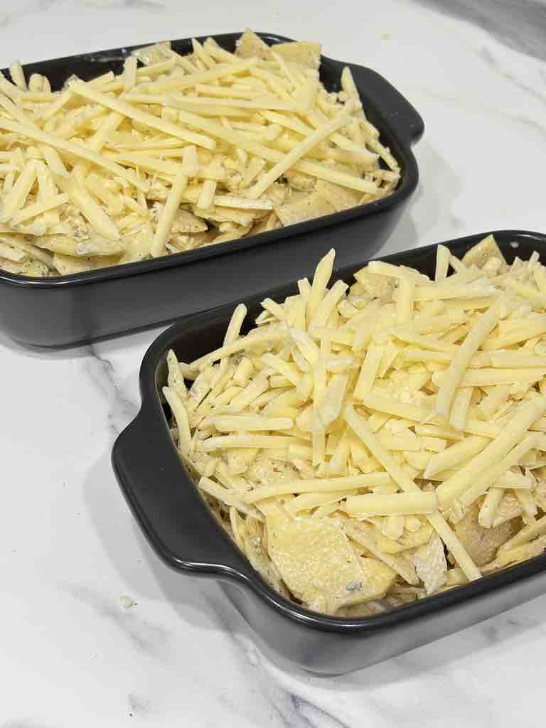 Two black baking dishes filled with creamy potato slices and topped generously with shredded cheese, prepped for baking into a golden potato gratin.