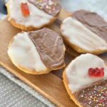 Top view of black and white cookies with vibrant decorations including chocolate glaze, white icing, rainbow nonpareils, and gummy bears, served on a rustic wooden board.