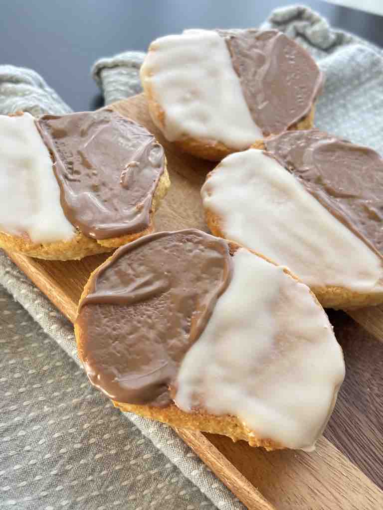 Amerikaner cookies in their traditional form, iced with smooth chocolate and white sugar glaze, no extra toppings, served on a wooden board with a neutral napkin.