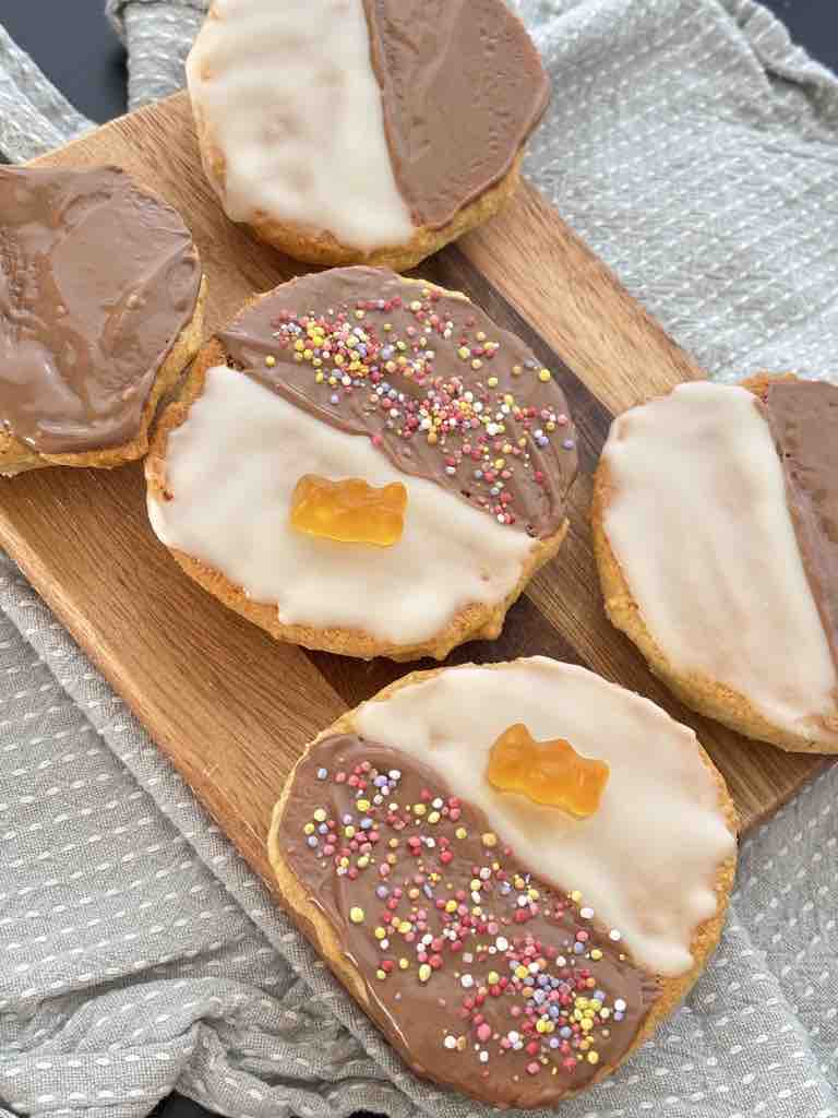 Assortment of decorated and traditional Amerikaner cookies, some with sprinkles and gummy bears, others with plain black and white icing, all on a wooden board with a cloth backdrop.