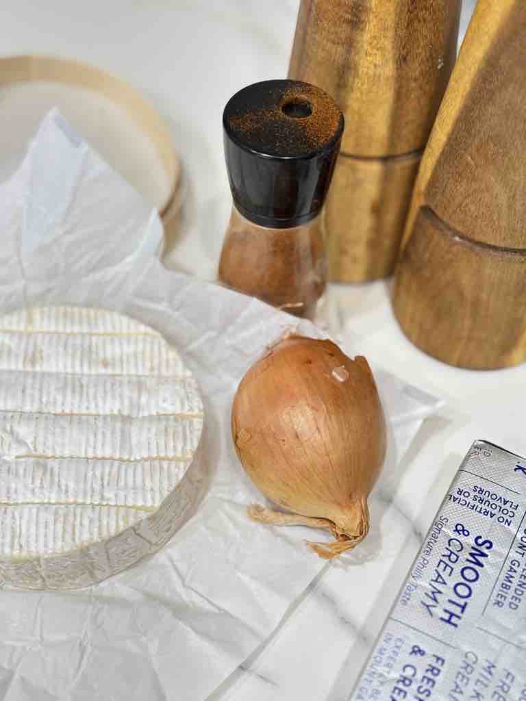 Ingredients for an obatzda recipe without beer, including Camembert cheese, onion, paprika, and cream cheese on a countertop.