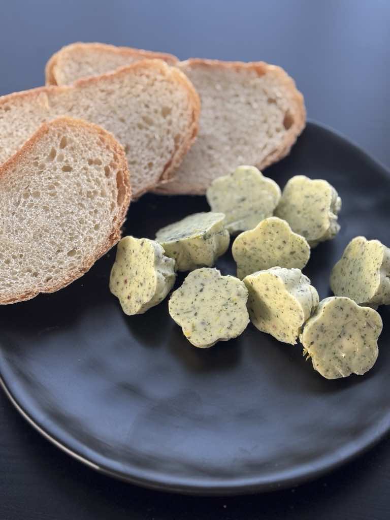 A black plate filled with rustic German bread slices and several frozen herb butter flowers, from a simple and old-school recipe.