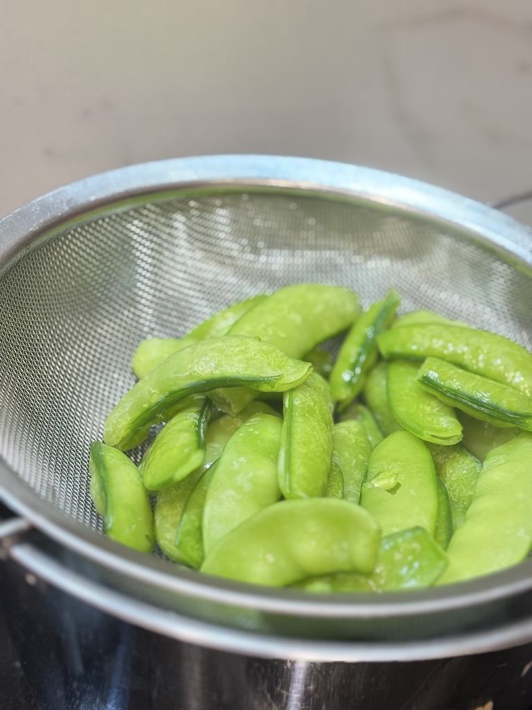 Sugar snap peas being steamed in a metal sieve over a pot. A key visual in the process of how to steam sugar snap peas to keep them crisp and vibrant.