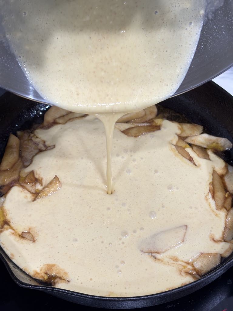 Pancake batter covering apple slices in a hot cast iron pan — the first step to a golden, oven-baked German pancake.