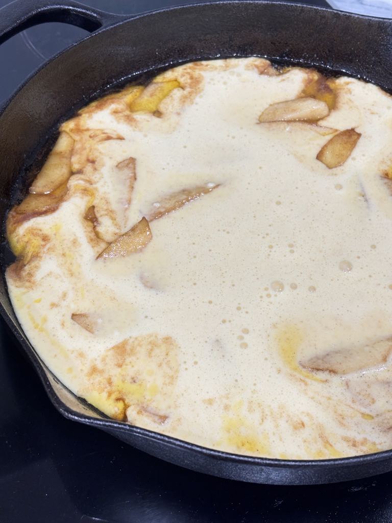 A cast-iron pan filled with apple-studded batter, ready to go into a preheated oven for a fluffy, puffy pancake.